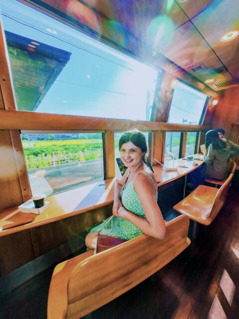 A smiling woman in green dress seated at the wooden counter of the Yufuin no Mori sightseeing train in Kyushu, Japan, enjoying the bright countryside views through the large panoramic windows that make the journey scenic and relaxing.