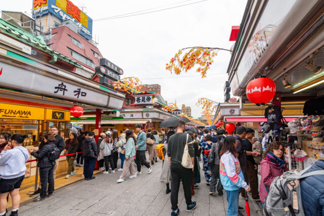 Crowds of tourists and locals shopping at Nakamise Street in Asakusa Tokyo, a lively shopping street leading to Sensoji Temple, decorated with red lanterns and autumn leaves.