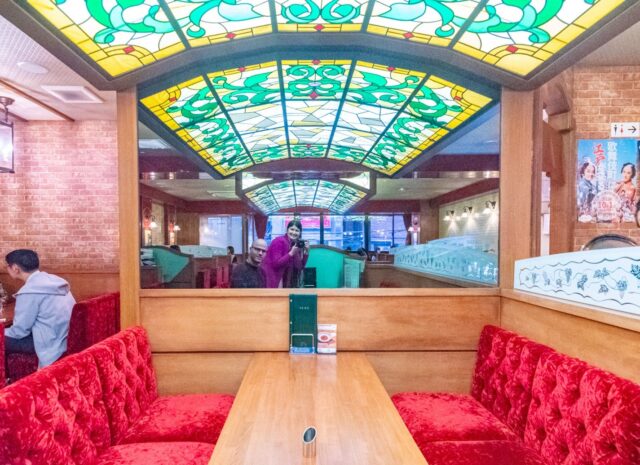 Interior of Coffee Seibu in Shinjuku Tokyo, featuring red velvet seating, wooden tables, and a stained-glass ceiling. A large mirror reflects two people, one holding a camera, capturing the scene. An example of traditional kissaten
