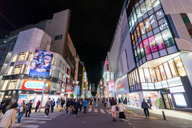 Bustling Ikebukuro Sunshine Street in Tokyo at night, filled with neon signs, anime billboards, arcades, shops, and crowds of people enjoying the nightlife.