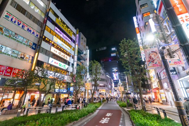 Neon-lit street in Shinjuku Kabukicho, Tokyo at night, with colorful billboards, restaurants, shops, and crowds of people walking under the bright city lights.
