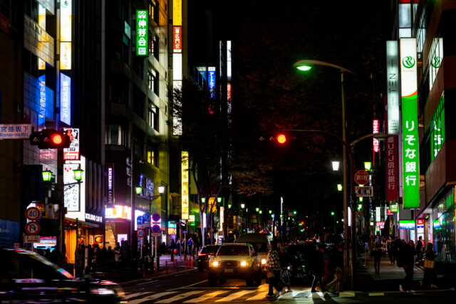 Busy Shinjuku street at night in Tokyo with neon signs, glowing billboards, cars at a red light, and pedestrians crossing under the city’s vibrant nightlife atmosphere.