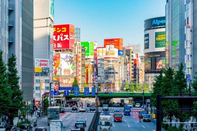 Busy Shinjuku Yasukuni Dori street in Tokyo during daytime, with colorful billboards, skyscrapers, traffic, and the iconic Yunika Vision screen in the background.