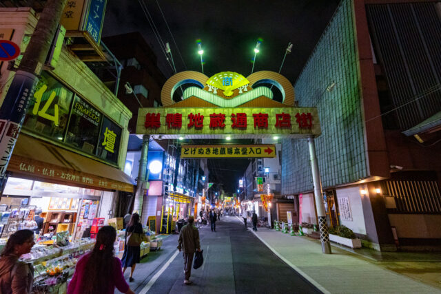 Entrance to Sugamo Jizo Dori Shopping Street in Tokyo at night, with illuminated sign, shops, and people walking along the bustling street.