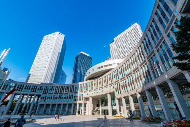 Wide-angle view of the Tokyo Metropolitan Government Building in Shinjuku, Tokyo, with its modern curved facade and towering skyscrapers under a bright blue sky.