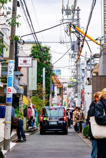 Busy street in Shimokitazawa Tokyo with pedestrians, boutique shops, overhead power lines, and a black taxi car driving through the narrow alley in a trendy shopping district.