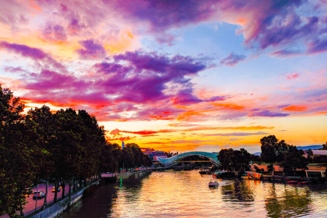 Vibrant sunset over the Mtkvari River with the illuminated Peace Bridge and colorful sky in Tbilisi, Georgia.