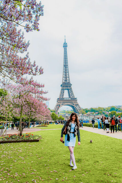 Woman with long dark hair in a light blue dress walking on the Champ de Mars lawn with the Eiffel Tower and spring blossoms.