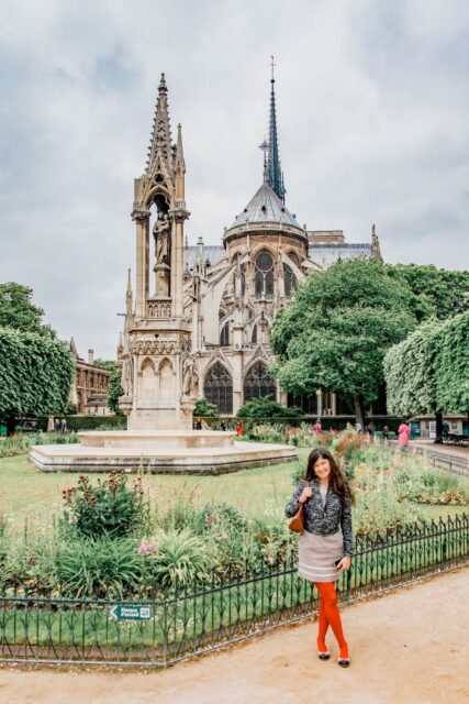Rear view of Notre-Dame with the Fountain of the Virgin in Square Jean-XXIII; woman with long hair, creamy dress and orange tights in the foreground.
