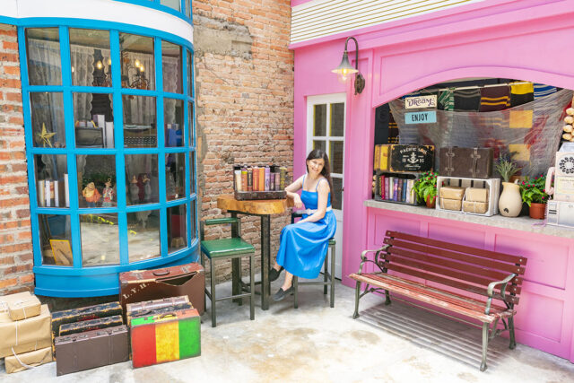 Woman with long dark hair in a bright blue dress by a pink storefront with a teal bay window and vintage suitcases in a cafe in Ipoh Malaysia.