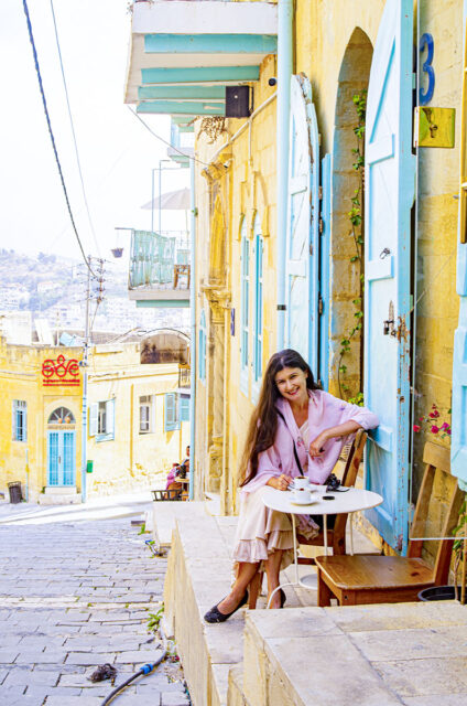 Woman with long dark hair in a pink dress enjoying coffee on a sunlit stone street with turquoise shutters.
