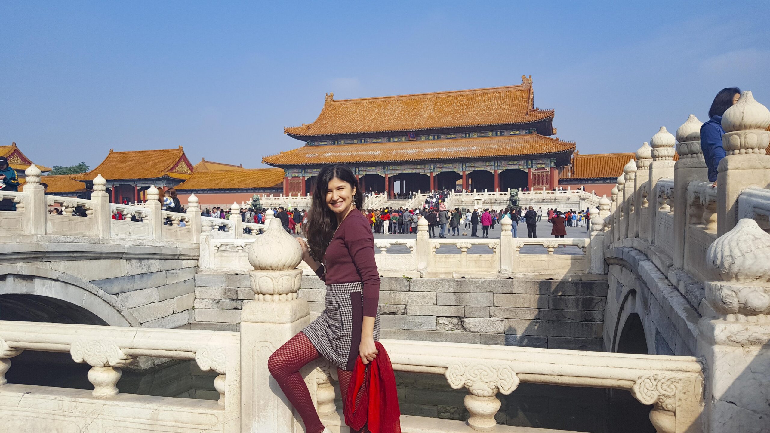 Ket, a smiling girl with long dark hair, wearing a maroon cardigan, checkered skirt, and red fishnet tights, posing on a white stone bridge in the Forbidden City, Beijing, China, with the grand imperial hall and golden roofs in the background on a sunny day.
