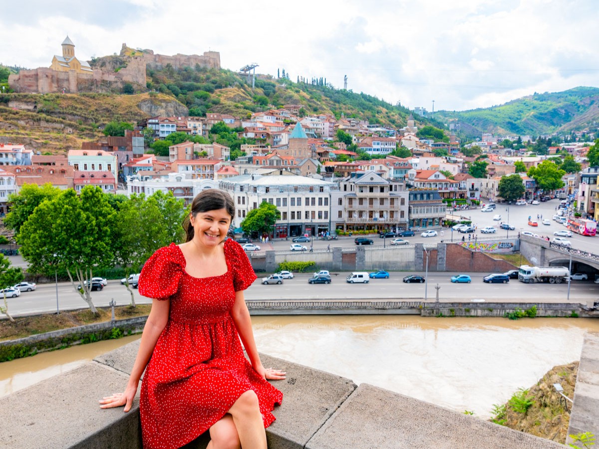 Woman in a red dress sitting by the Mtkvari River with a panoramic view of Tbilisi’s old town, Narikala Fortress, and historic churches in the background. Photo taken from Metekhi chutch