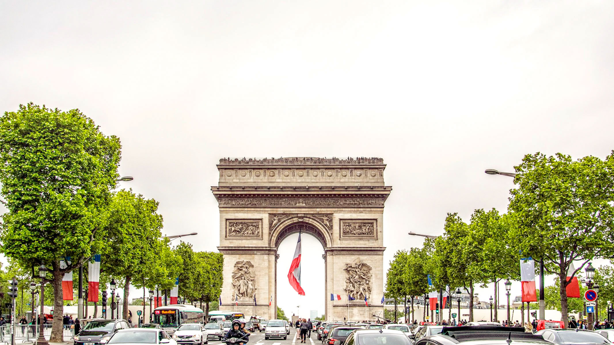 Arc de Triomphe centered on the Champs-Élysées in Paris, framed by plane trees and French flags, with city traffic under an overcast sky.