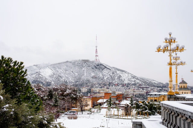 Snow-covered Mtatsminda Hill with the Tbilisi TV Tower overlooking the city skyline and golden street lamps in Tbilisi, Georgia.